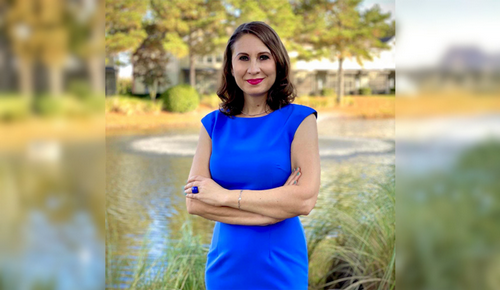 Woman in a bright blue dress stands with arms crossed by a pond, with trees and a building blurred in the background.