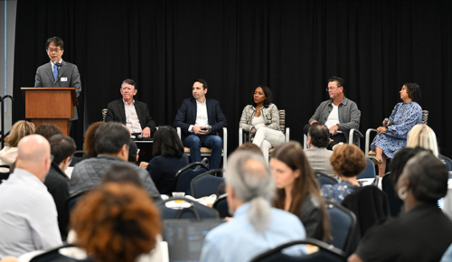 Sport industry professionals sit on stage for panel presentation next to standing Dr. Loh