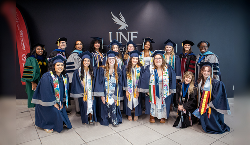 Group of graduates in caps and gowns posing with faculty in academic regalia in front of a wall displaying the UNF logo.