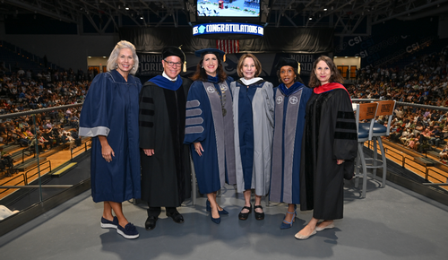 Five faculty members in academic regalia pose on a stage at a graduation ceremony, with a packed arena and a “CONGRATULATIONS” scoreboard in the background.
