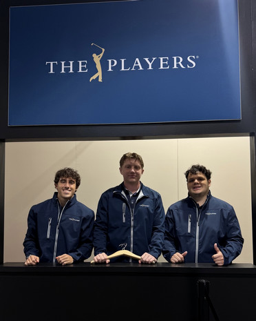Three people in navy jackets stand behind a check-in counter beneath a “THE PLAYERS” sign.