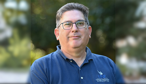 Outdoor head-and-shoulders portrait of a man wearing glasses and a navy UNF polo shirt, with a softly blurred green background.