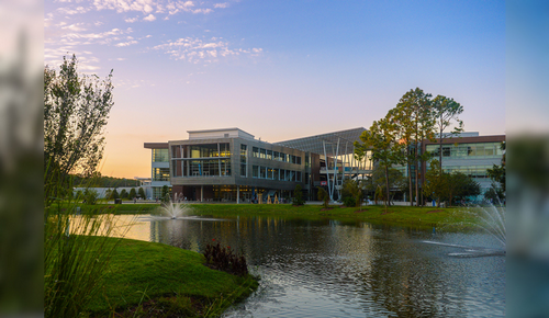 Modern office building with glass windows beside a pond with fountain sprays, set in landscaped greenery at sunset.