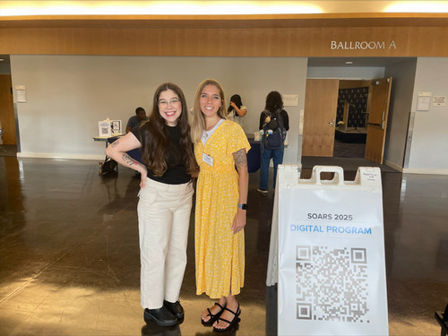 Two women smile in front of a SOARS 2025 digital program sign.