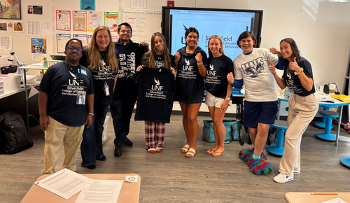 Group of eight smiling people in a classroom holding UNF-branded shirts, standing in front of a large screen and classroom posters.