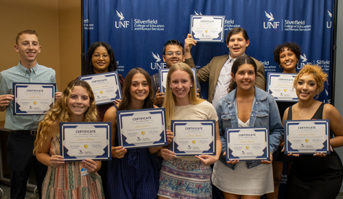 Group of smiling students holding certificates in front of a UNF Silverfield College of Education and Human Services backdrop.