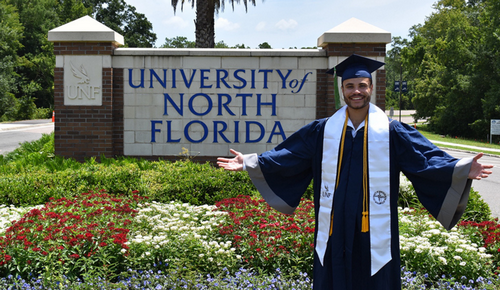 Michael Carrero standing in front of UNF entrance sign with cap and gown