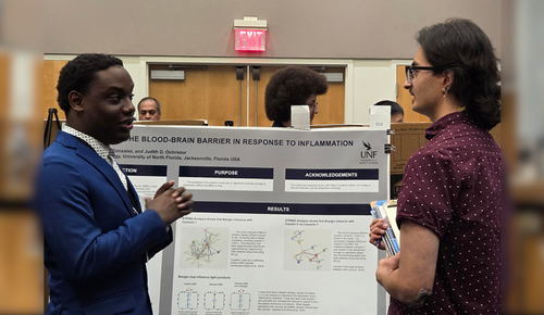 Two people talk in front of a scientific research poster at a conference, with the presenter gesturing while explaining the results.
