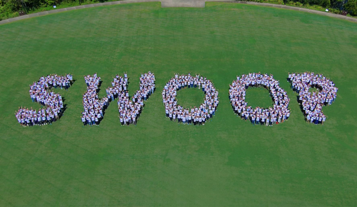 Aerial view of a large group of people standing on a grassy field arranged to spell “SWOOP.”