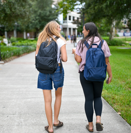Two UNF students with blue and black backpacks walking and talking outside 