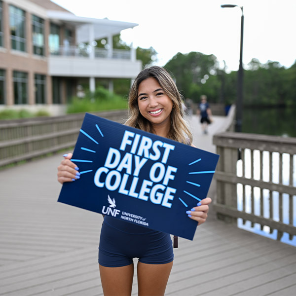 student standing on a bridge and smiling while holding a first day of college sign