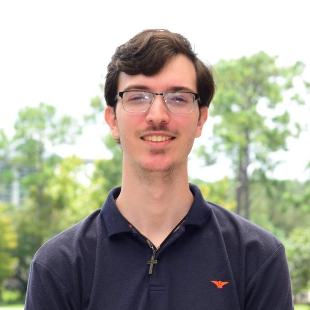 Daniel wears a blue shirt and stands in front of trees