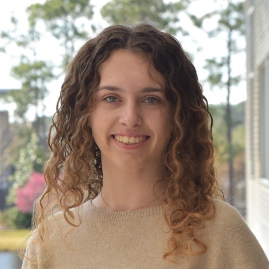 Makayla has curly hair and is wearing a tan sweater.