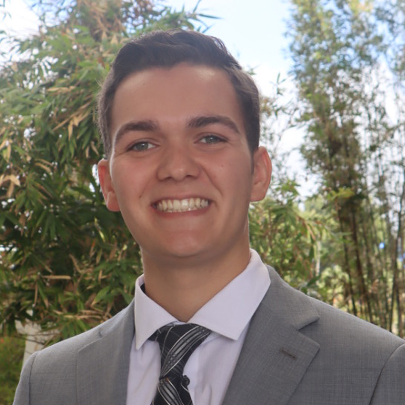 Pablo in a grey suit and black tie standing in front of bamboo