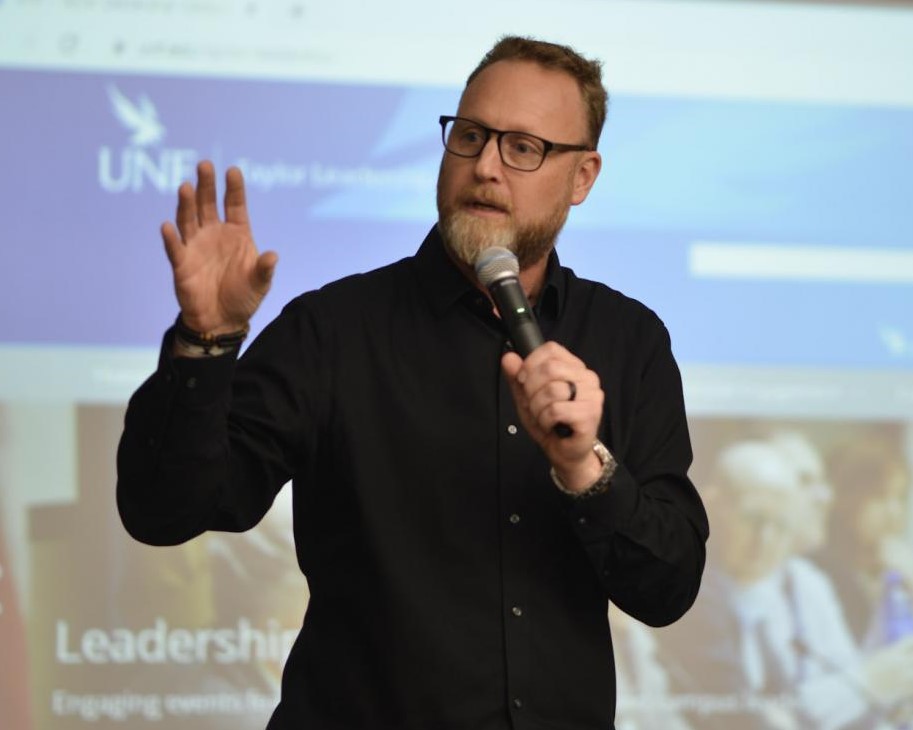 Speaker Jonathan Catherman speaking into a microphone in front of a projector screen at a TLI event