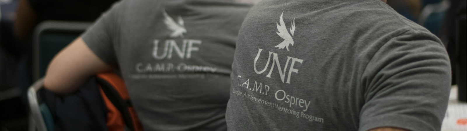 View of two students seated with only the back of their gray shirts visible with the UNF CAMP Osprey logo 
