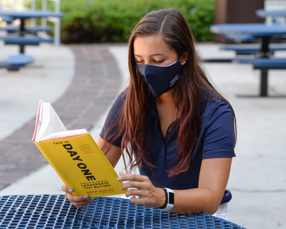 Female student wearing a mask and reading a book at an outdoor table in Alumni Square