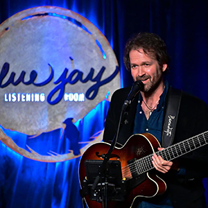 Photo of Taylor Roberts holding a guitar talking into microphone