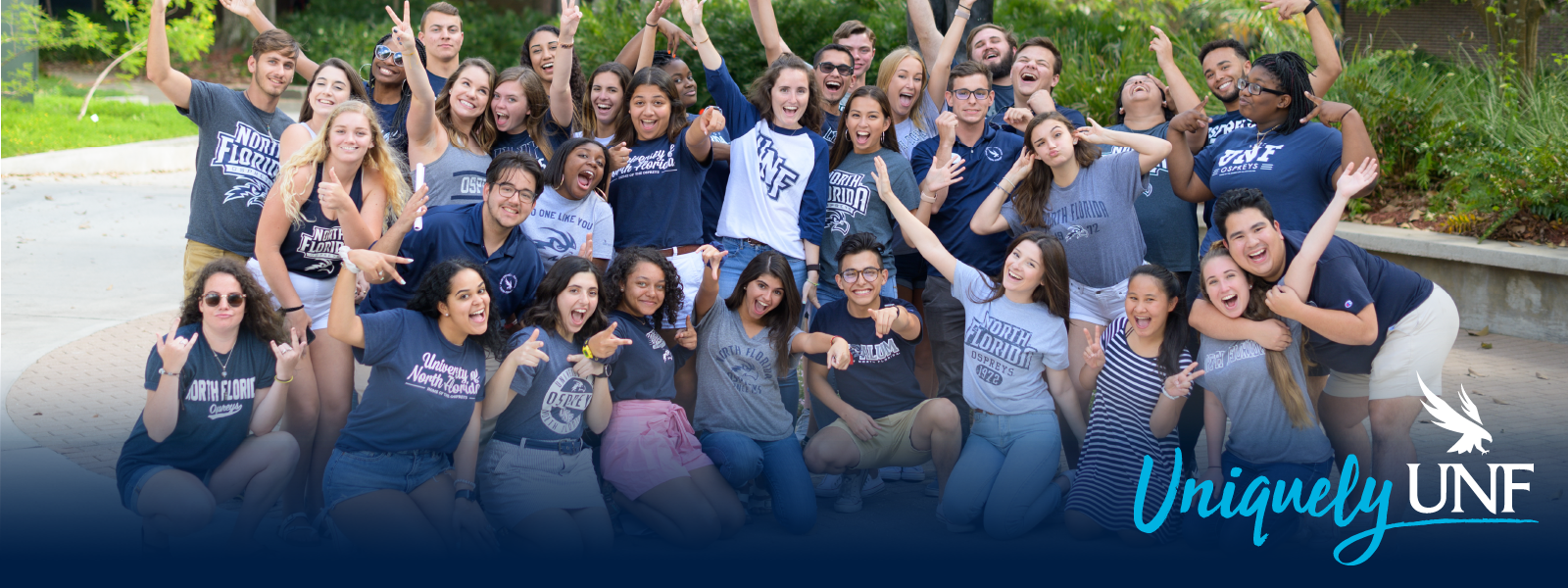 Large group of UNF students in UNF t-shirts all smiling largely with their hands up and the Uniquely UNF logo