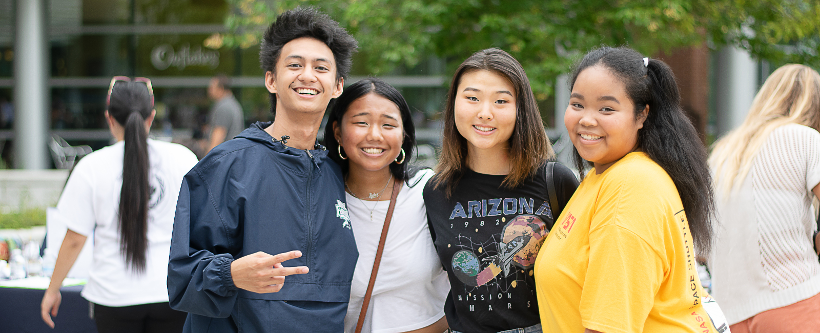 Four students outside the student union all huddled together and smiling and holding up a peace sign