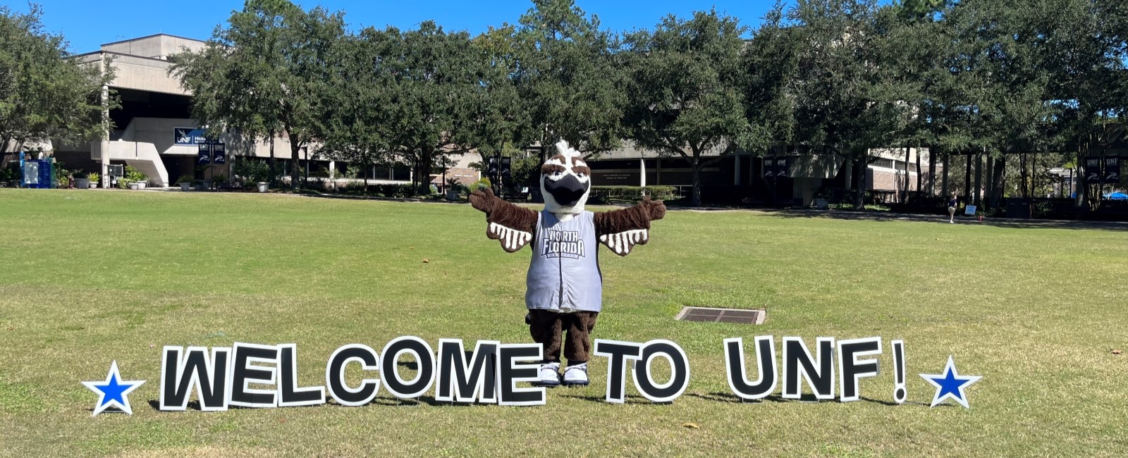 Ozzie Osprey standing on the green in front a sign that says welcome to UNF