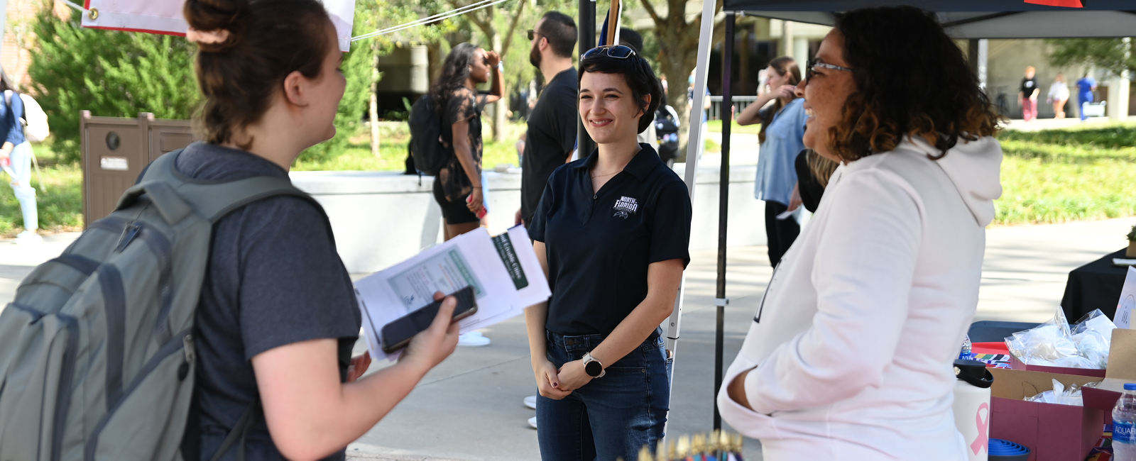 Female UNF employee wearing a UNF polo standing outside by a tent smiling at a student in conversation