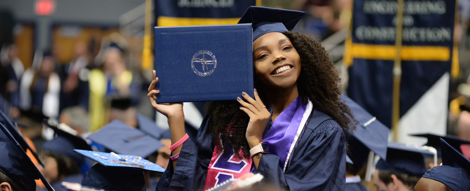 Female student at a UNF graduation in a cap and gown holding a diploma cover up and smiling