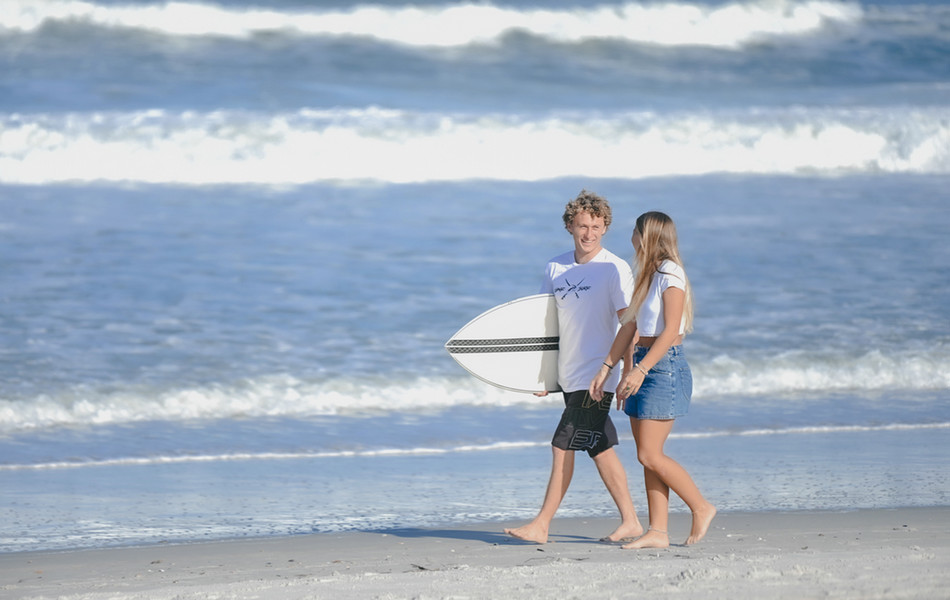2 students walking on the beach and smiling with one carrying a surfboard